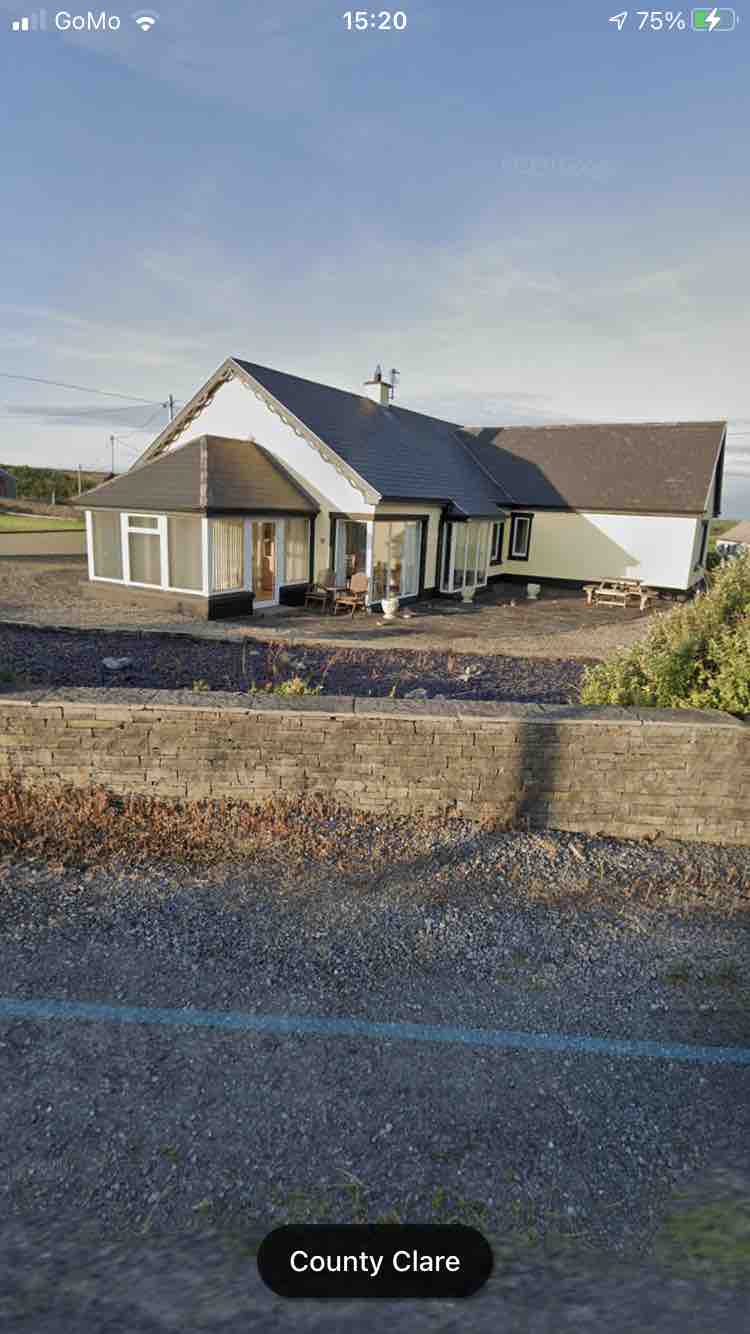 A cottage lodge with a distinctive design features large windows that provide ample natural light. Surrounded by greenery, a spacious patio is visible, offering outdoor seating for relaxation. The structure displays a blend of traditional and modern architecture, set against a serene rural backdrop.