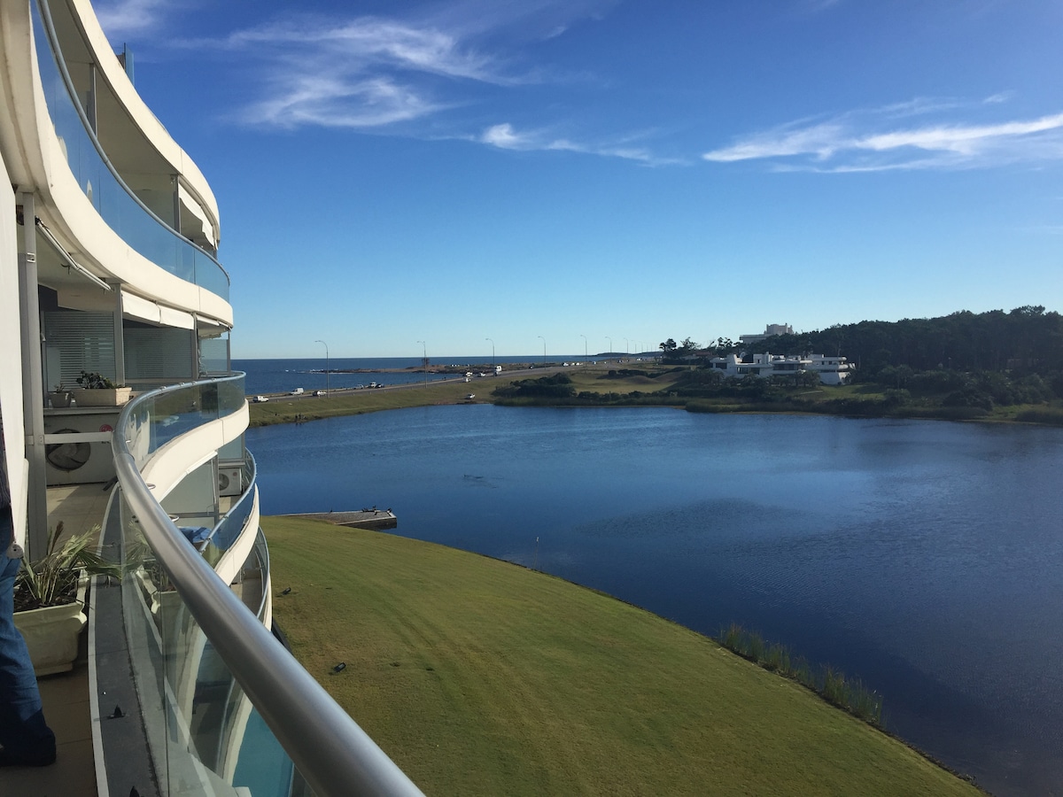 A curved balcony view captures the expansive landscape, featuring a serene lagoon bordered by lush green grass and a clear blue sky. The distant shoreline provides a backdrop, while the gentle ripples on the water's surface reflect the sunlight.