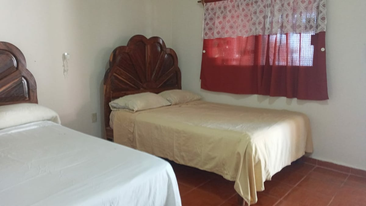 A bedroom features two beds with wooden headboards, one made up with a white coverlet and the other with a cream blanket. Natural light enters through a red and white patterned curtain covering a window.