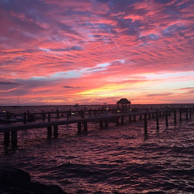 A vibrant sunset casts shades of pink and orange across the sky, reflecting on the gentle waves of the water. A wooden pier extends into the bay, leading toward a thatched-roof gazebo, creating a picturesque scene of the waterfront.