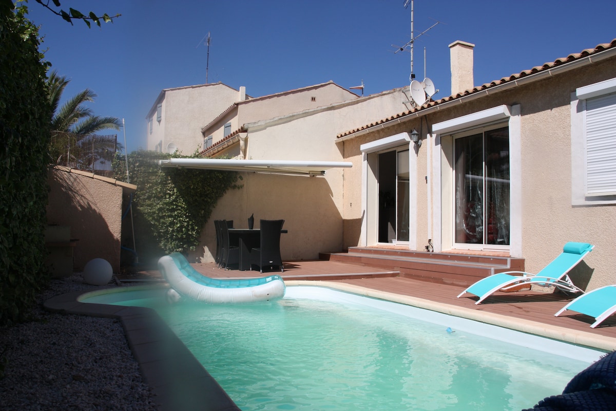 A small private pool is surrounded by a wooden deck, with two lounge chairs positioned nearby. A retractable awning provides shade over a seating area. The house features large glass doors offering a view of the pool area, framed by lush greenery.