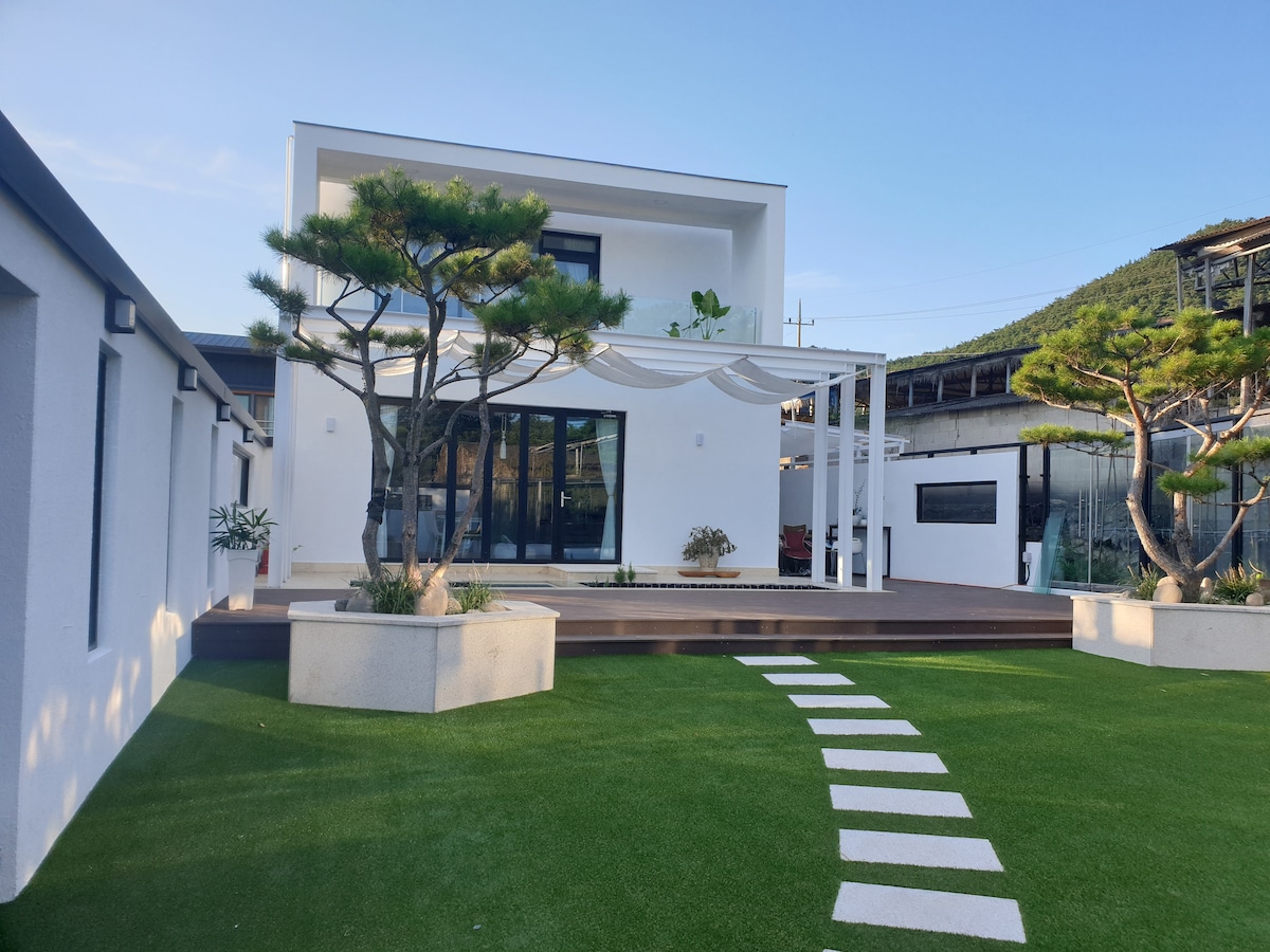 The exterior features a modern two-story building surrounded by a landscaped yard. A spacious deck with a pergola is visible, offering a shaded area. Neatly arranged stepping stones lead through green grass, with well-maintained trees adding natural elements to the outdoor setting.