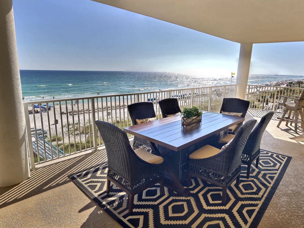 A spacious deck features a large dining table surrounded by eight wicker chairs, offering unobstructed views of the ocean. The sun reflects off the water, creating a sparkling effect. A patterned outdoor rug enhances the setting, inviting guests to enjoy meals in the open air.