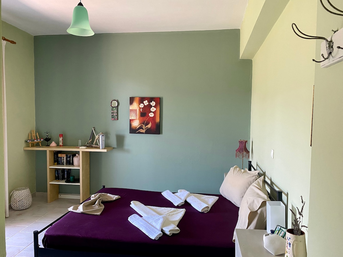 A serene bedroom is presented with calming green walls. A spacious bed is neatly arranged with two white towels. A wooden shelf holds various items, while a piece of wall art adds a touch of color to the space. Natural light enhances the welcoming ambiance.
