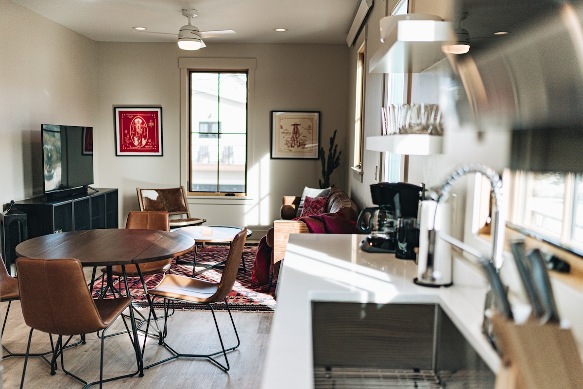 An open living area features a circular dining table surrounded by modern chairs. A comfortable sitting area showcases a plush sofa and decorative art pieces on the walls. A well-equipped kitchen with sleek fixtures and ample counter space is visible, enhancing the inviting atmosphere.