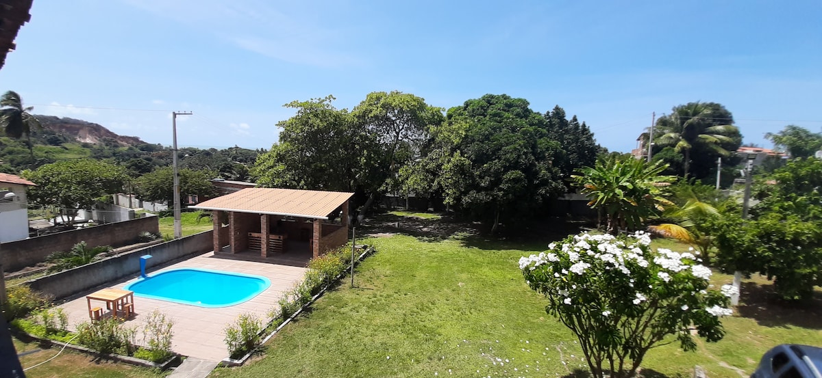 An expansive outdoor area features a clear blue swimming pool surrounded by lush greenery. A wooden gazebo is visible nearby, offering shaded seating. Fruit trees and flowering bushes enhance the natural environment under a clear blue sky.