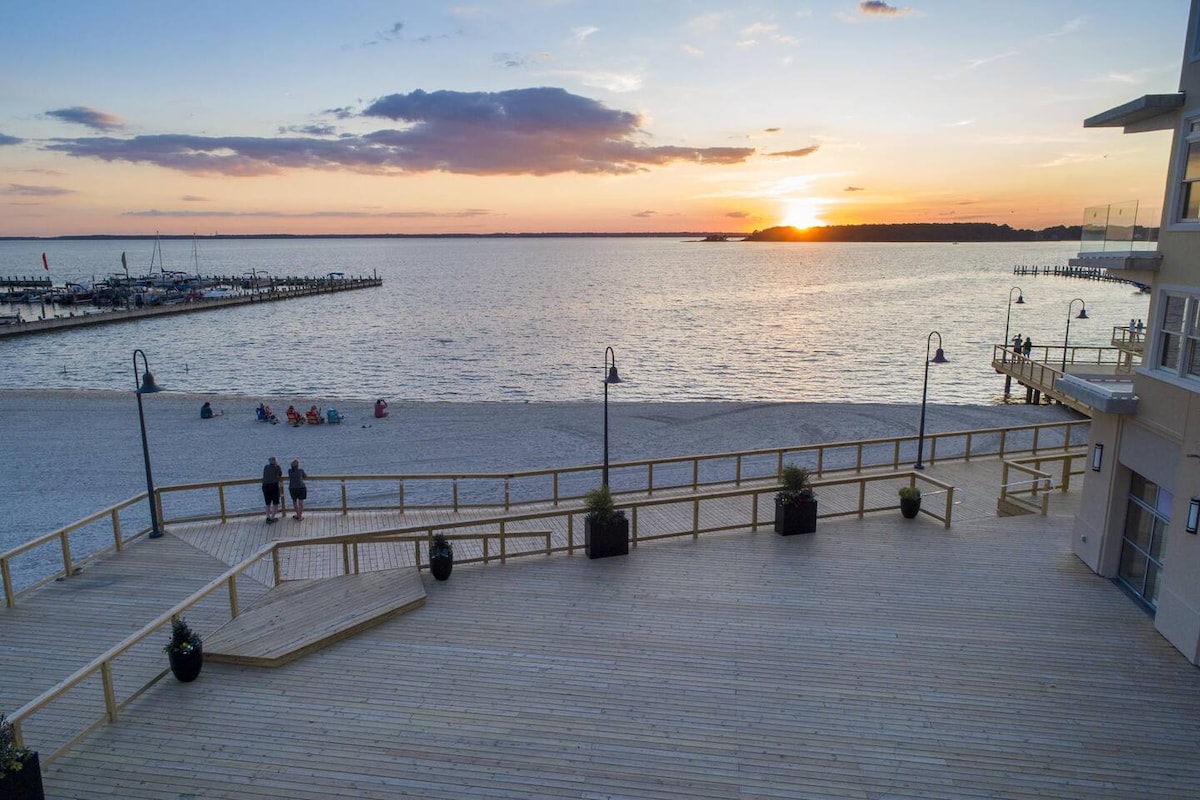 A wooden boardwalk leads to a sandy beach, where visitors can be seen enjoying the sunset over the calm bay waters. Lamp posts line the path, providing gentle light as the sun sets behind the horizon, creating a serene atmosphere.