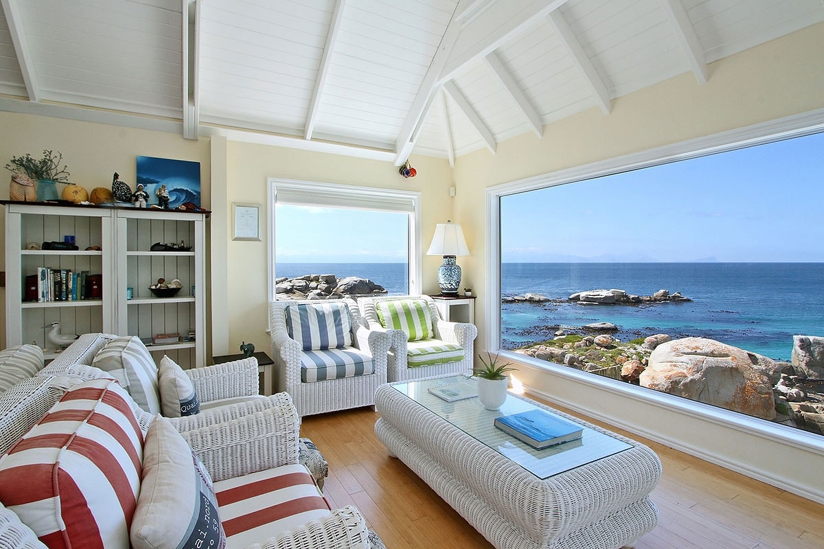 A light-filled living area is highlighted by large windows offering unobstructed ocean views. Comfortable seating consists of a striped sofa and armchairs arranged around a glass coffee table. Natural wood flooring complements the bright, airy ambiance enhanced by the coastal setting.