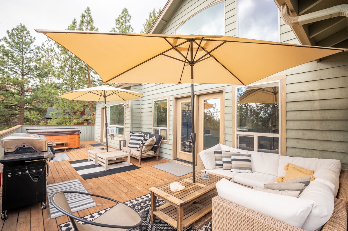 An outdoor patio is equipped with two umbrella-shaded seating areas, featuring a sectional sofa and wooden coffee table. A gas grill is positioned nearby, and a private hot tub is visible in the background, surrounded by trees and natural scenery.