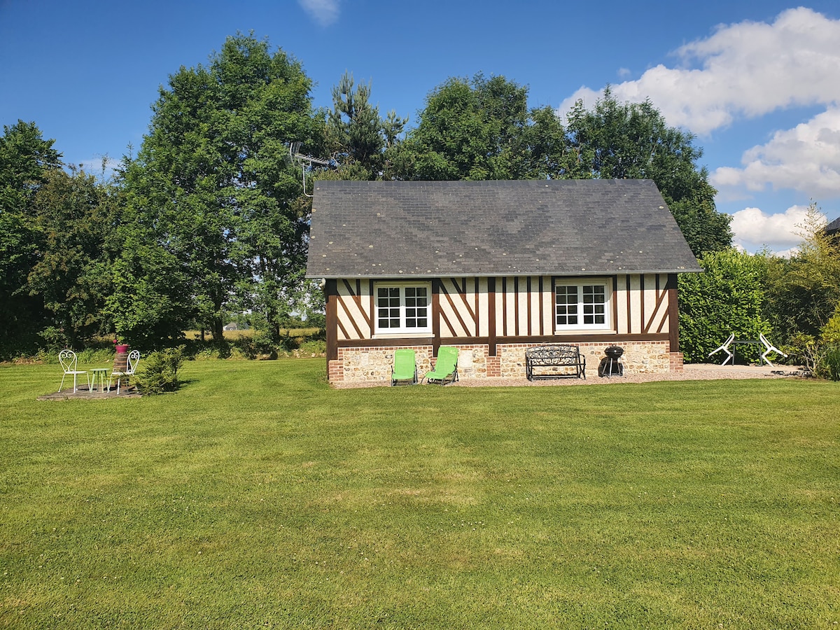 A charming countryside cottage is set within a lush green lawn, surrounded by trees. The exterior features traditional half-timbered design, and large windows allow natural light to fill the space. Chairs are placed in the garden for relaxation, creating an inviting outdoor area.