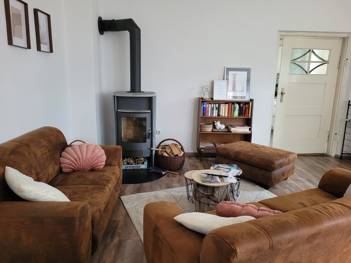 A cozy living area features two brown couches and a compact coffee table on a light-colored rug. A wood-burning stove adds warmth, while a bookshelf displays a collection of books. A basket with logs is positioned nearby, offering convenient access.