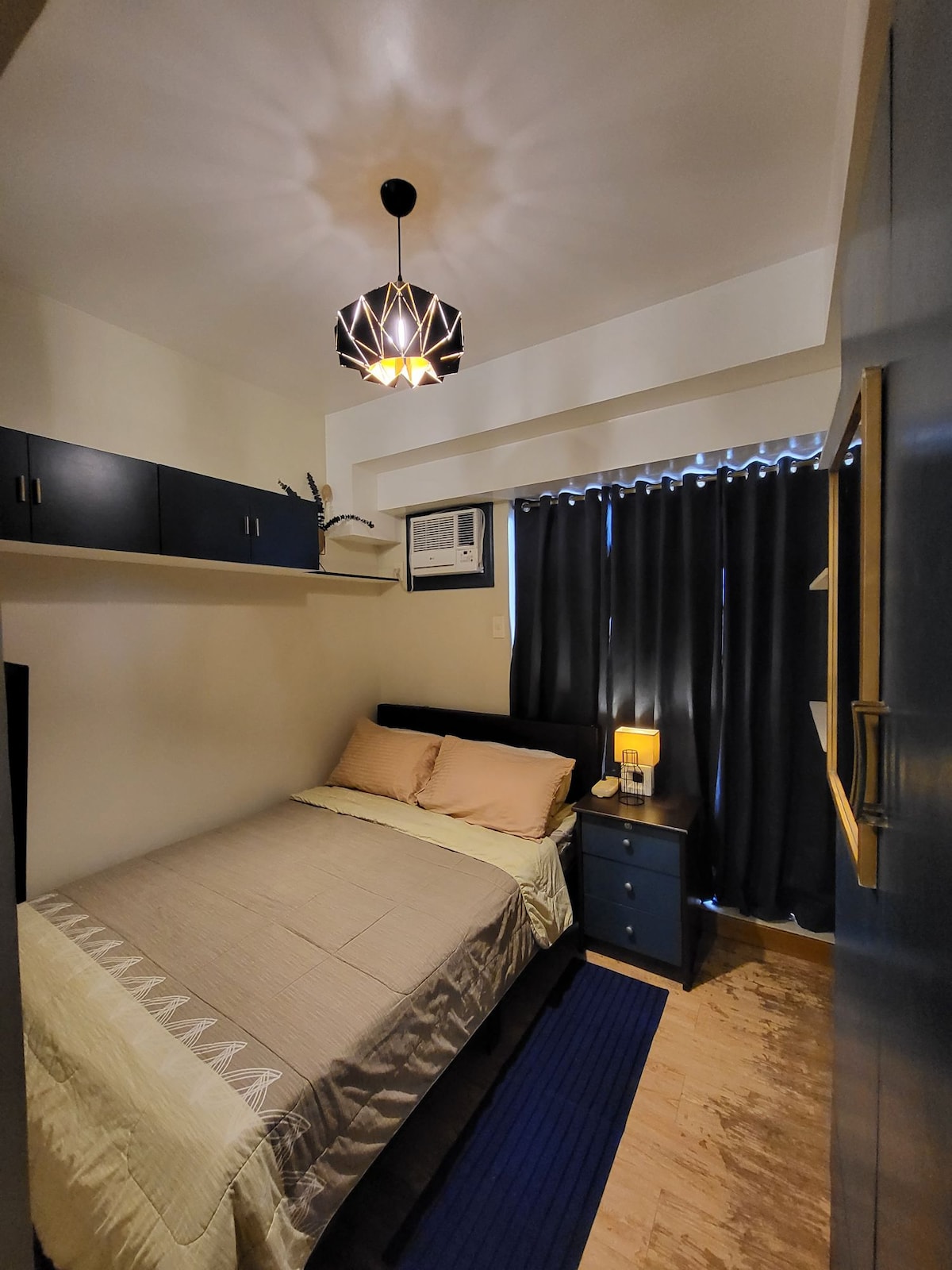 A compact bedroom is furnished with a bed covered in a light-colored blanket. A bedside table holds a lamp, and shelves are installed above for additional storage. Natural light filters through the dark curtains, while an air conditioning unit is mounted near the window.
