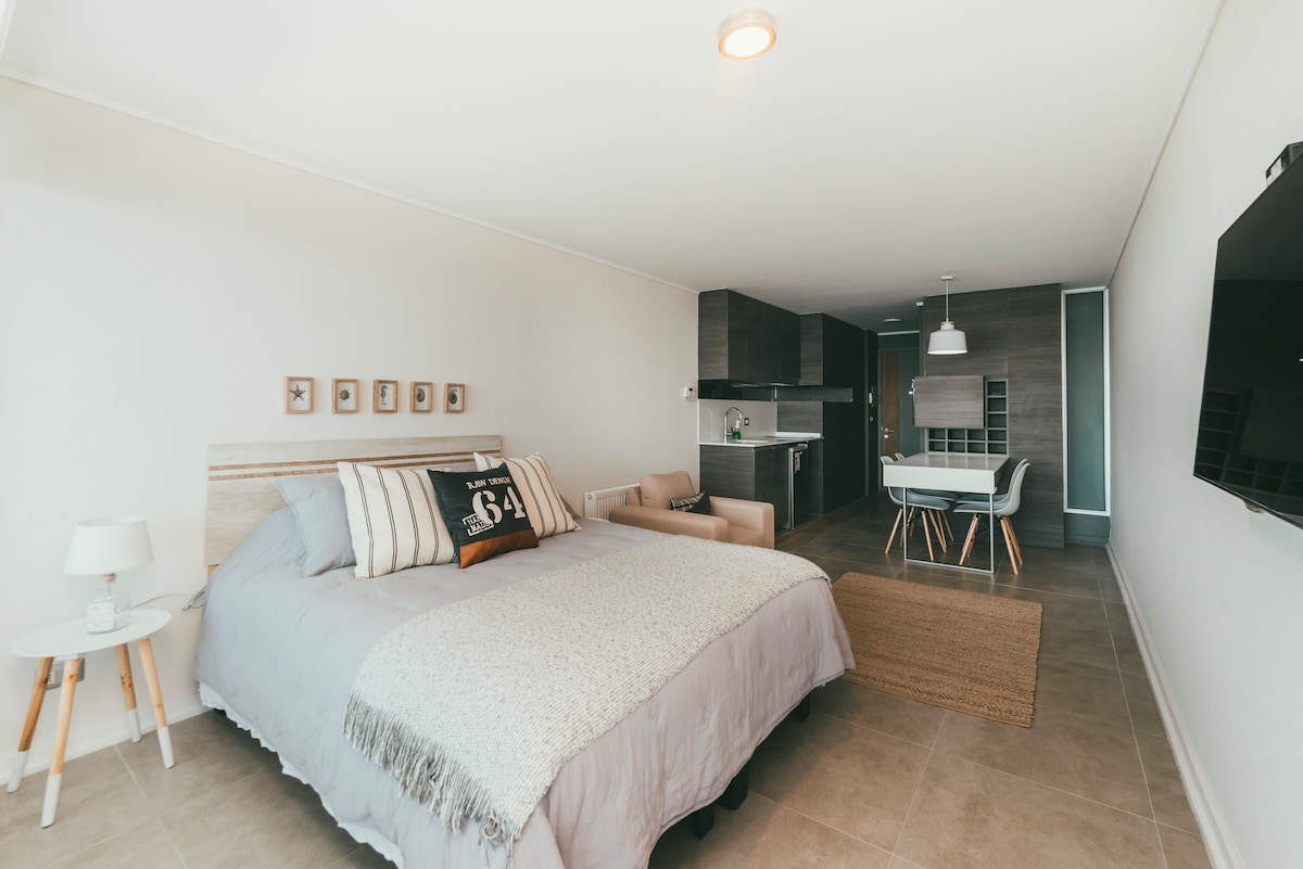 A spacious studio apartment features a comfortable bed with light bedding and decorative pillows. A modern dining area is visible in the background, complemented by a sleek kitchen setup. Soft lighting highlights the neutral color palette and stylish design throughout the room.