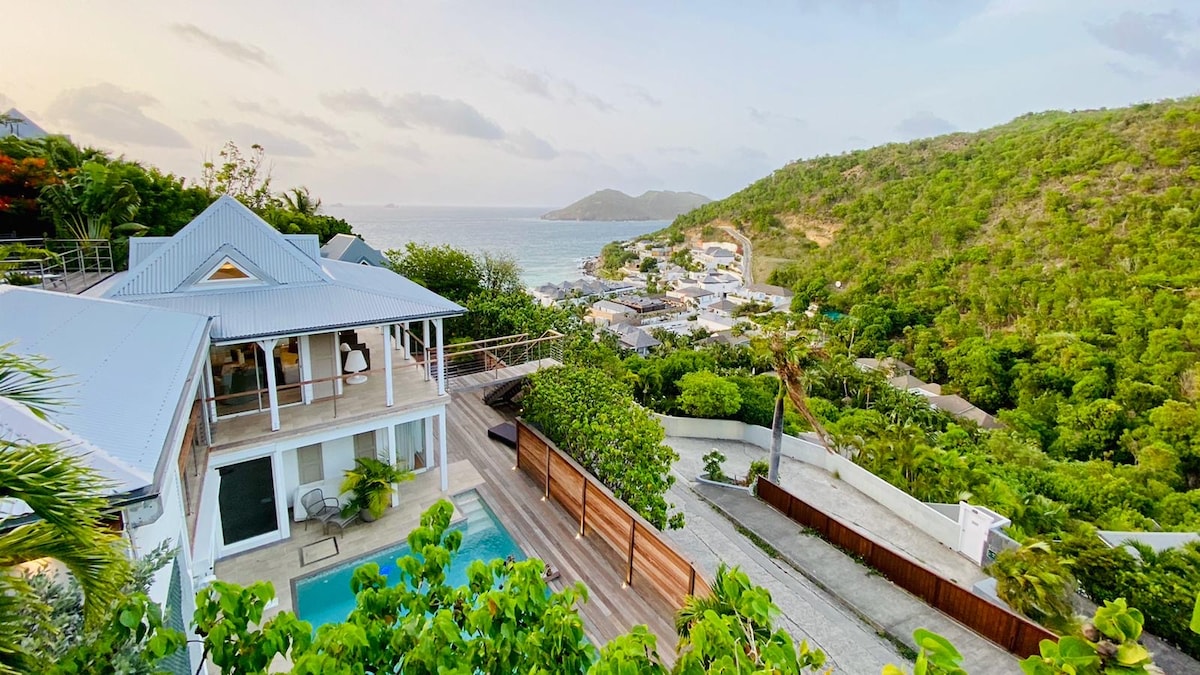 The villa is set against a verdant hillside, with a clear view of the coast and ocean in the distance. A private pool is visible in the foreground, surrounded by lush greenery. The modern architecture of the villa complements the scenic landscape.