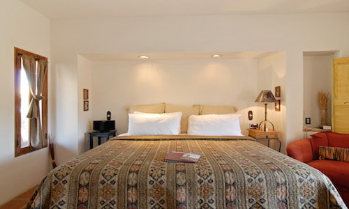 A spacious sleeping area features a large bed adorned with a patterned coverlet and multiple pillows. Natural light fills the room, highlighting the simple décor. A cozy seating area is positioned nearby, complemented by a lamp and wall accents.
