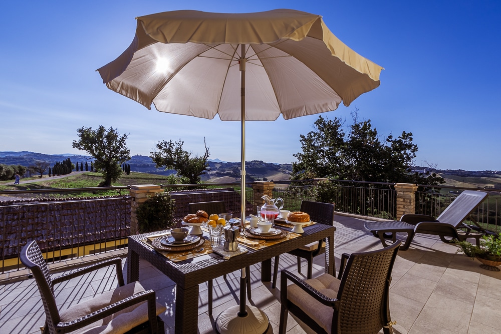 An outdoor dining area features a large umbrella providing shade over a stylish table set with dishes and pastries. A picturesque landscape of rolling hills is visible in the background, complemented by a sun loungers nearby, enhancing the relaxing atmosphere.
