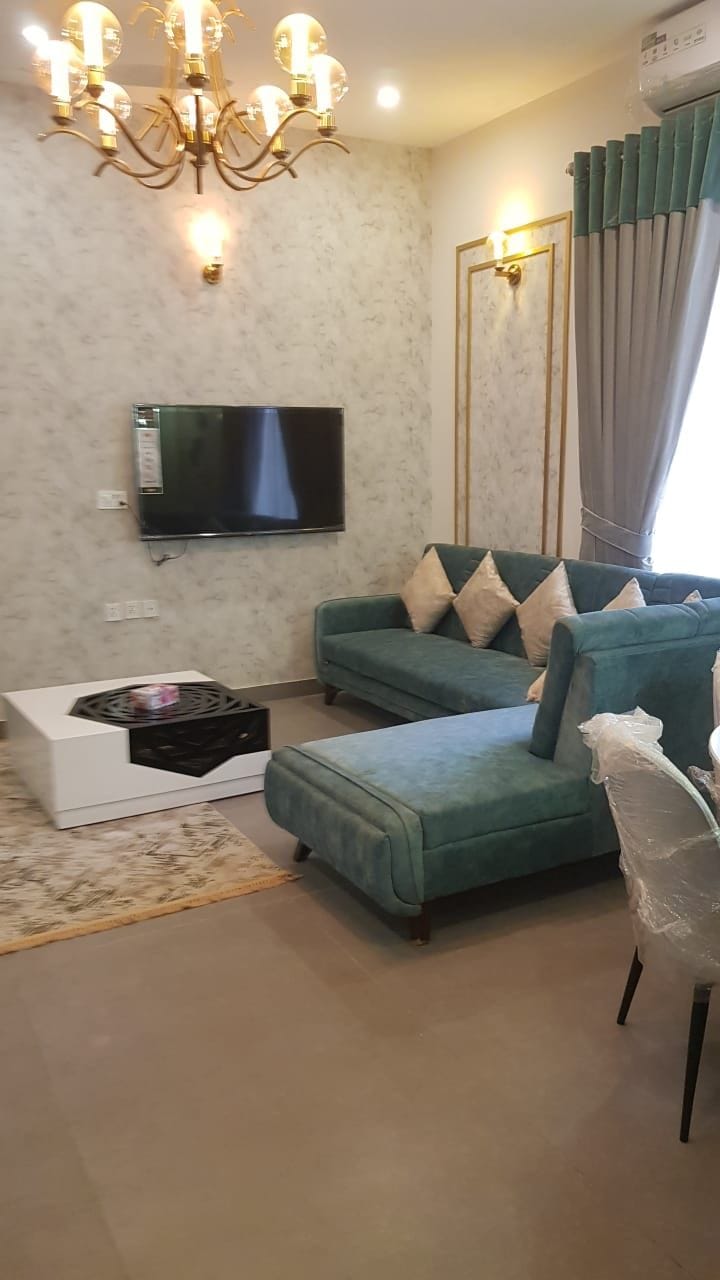 A well-furnished living area features a plush teal sofa with decorative cushions and a sleek, modern television mounted on the wall. A stylish white coffee table complements a textured area rug, while sheer curtains allow soft light to filter in through a window.