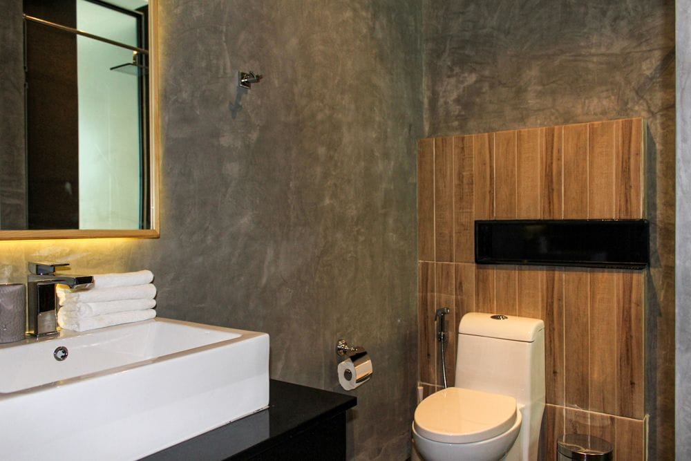 The bathroom features a modern design with a combination of textured grey walls and wooden accents. A rectangular sink is placed atop a dark cabinet, with neatly arranged white towels beside it. A contemporary toilet is located next to a black wall element, complemented by a mirror above the sink.