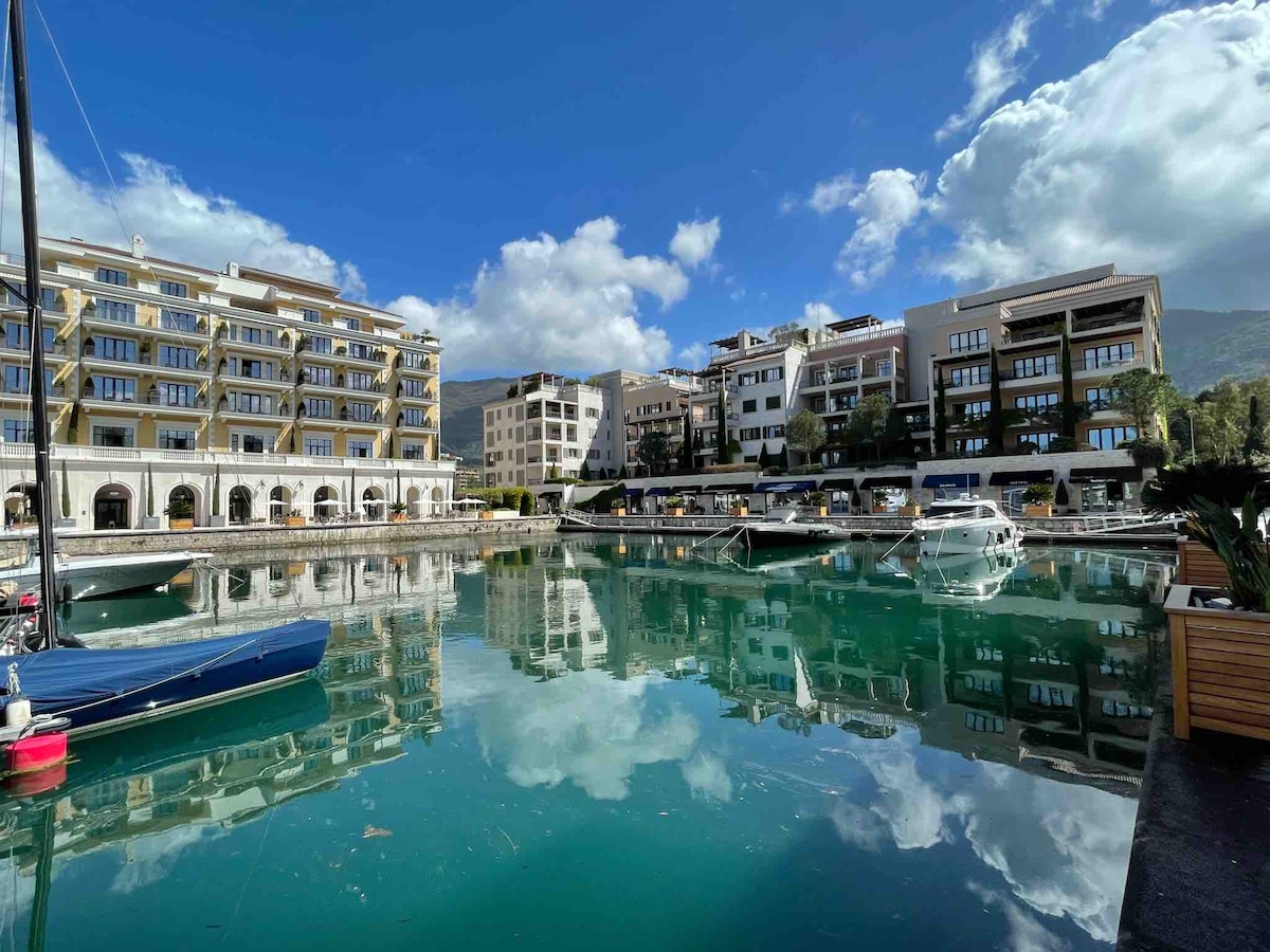 A serene marina scene is depicted, featuring boats gently floating on calm waters that reflect the surrounding buildings and clouds. Modern structures line the waterfront, providing a picturesque backdrop to the vibrant turquoise water.