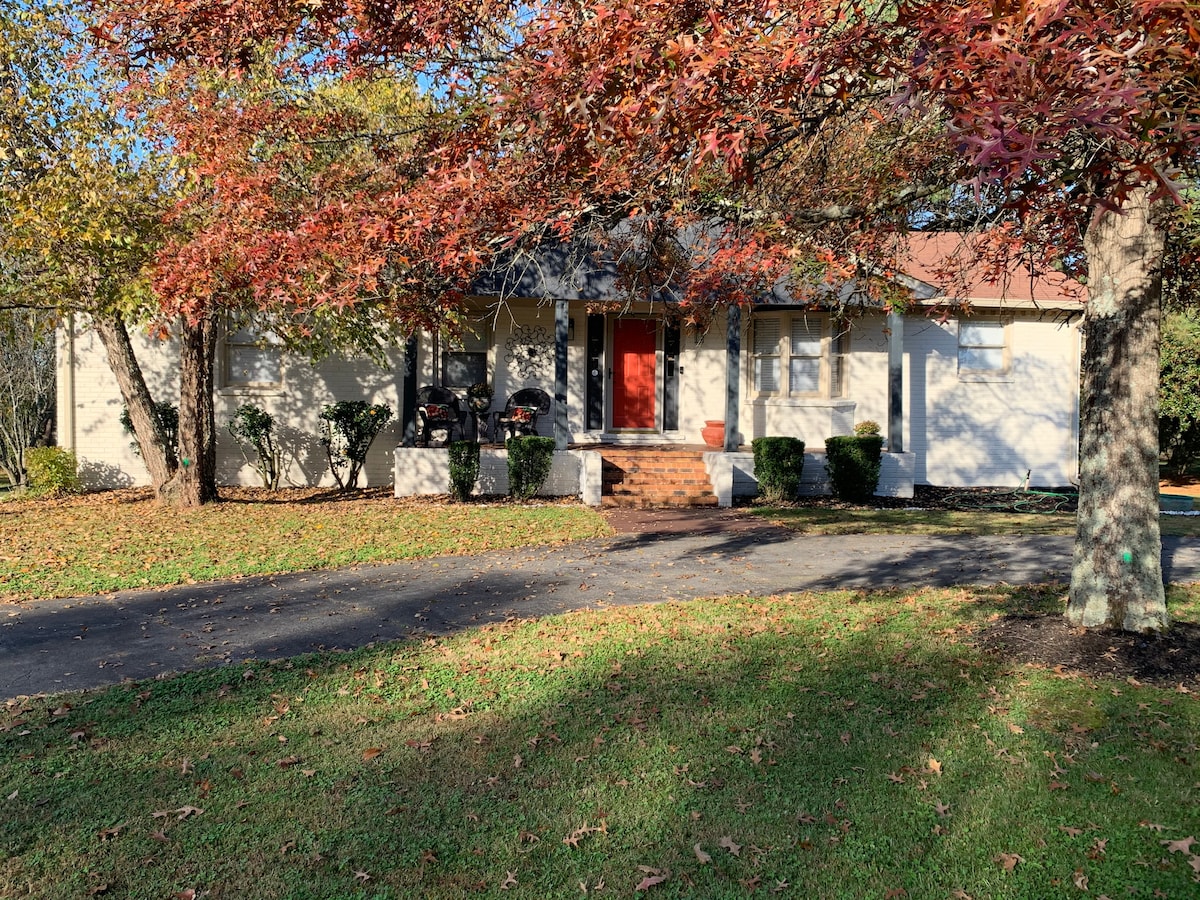 The exterior of the home is shown, featuring a welcoming red front door framed by steps and a porch. Surrounding trees display autumn leaves, and well-maintained shrubs line the pathway. A paved drive leads up to the home, contrasting with the lush green lawn.