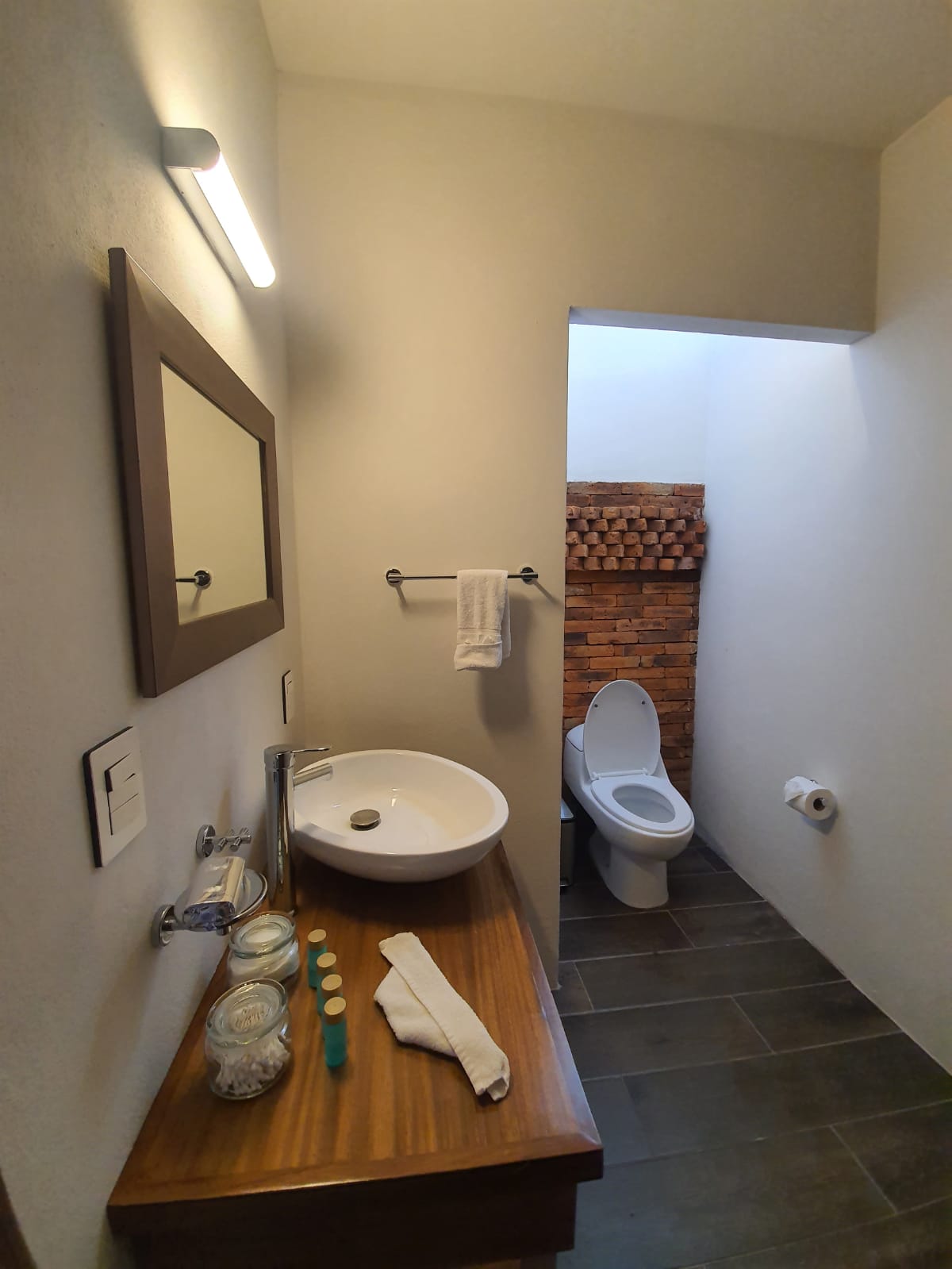 A bright bathroom features a minimalist design with a wooden vanity and a circular basin. A large mirror reflects natural light, while a discreet toilet is positioned at the rear. Textured brick accents add warmth to the space, enhancing the overall aesthetic.