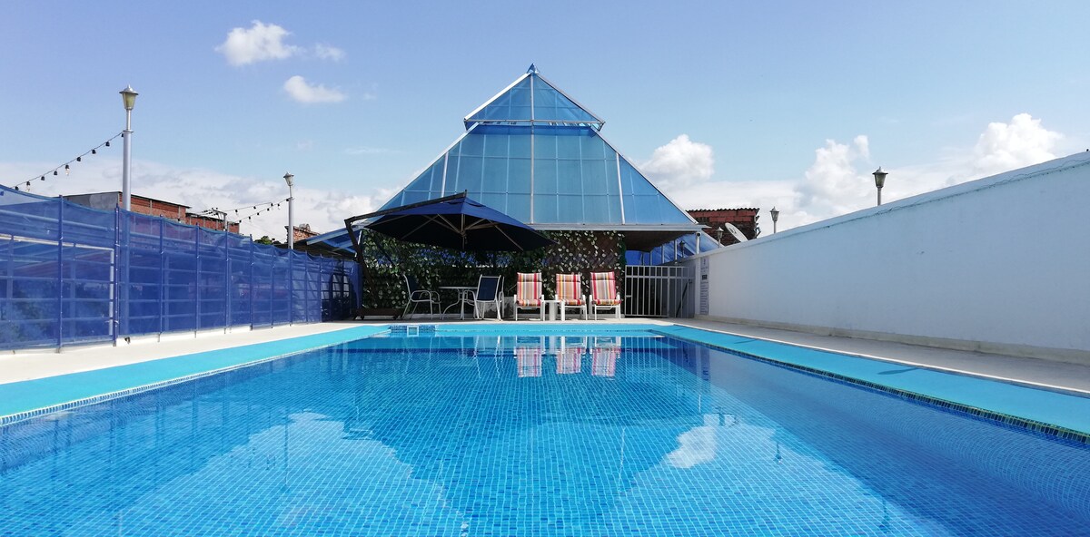 A large outdoor pool reflects the blue sky, surrounded by a white wall. A pyramid-shaped structure with a glass roof is visible in the background, accompanied by several lounge chairs under blue umbrellas, providing a spacious area for relaxation.