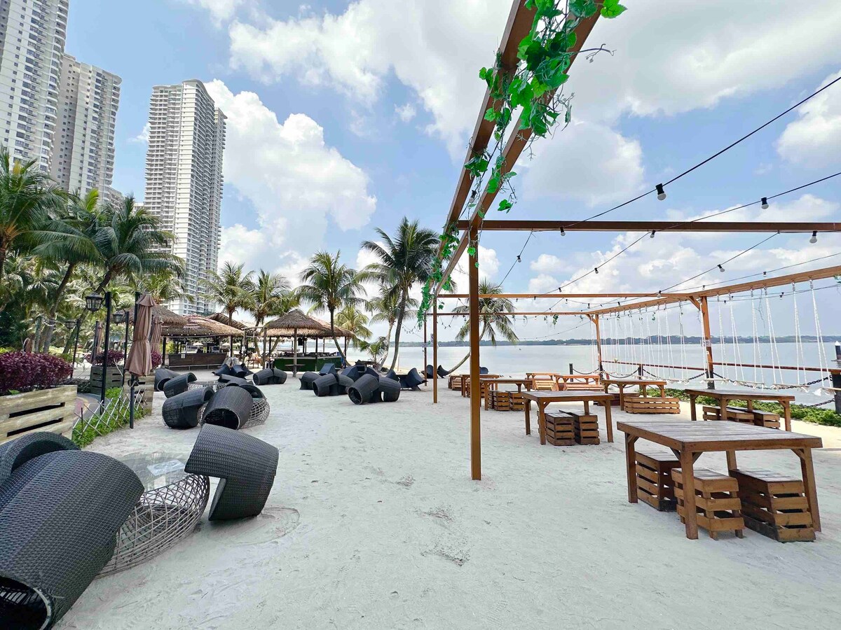 An outdoor seating area is presented, featuring several black lounge chairs arranged on sandy ground. Wooden tables are positioned throughout, surrounded by tropical palm trees. In the background, high-rise buildings rise against a cloudy sky, creating a relaxed coastal atmosphere.