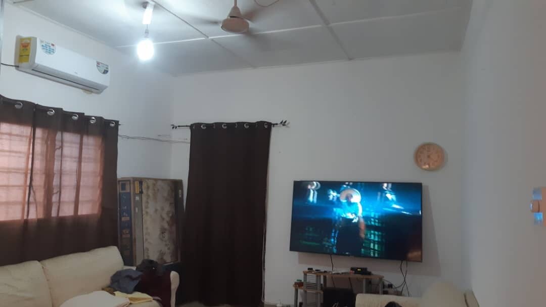 A living area features a large 65-inch 4K UHD Samsung TV mounted on the wall. An air-conditioning unit is visible above. Natural light filters through a window covered by brown curtains, while an inviting couch is positioned nearby, contributing to a relaxed environment.