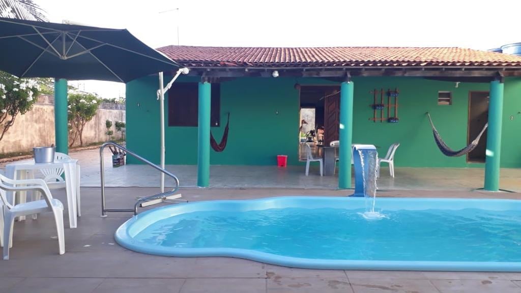 A tranquil outdoor area features a refreshing pool with a gentle waterfall, alongside white loungers and a large umbrella. The vibrant teal walls of the house provide a lively backdrop. Two hammocks are suspended nearby, inviting relaxation in the serene environment.