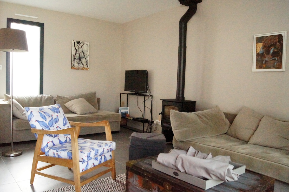 The living area features a cozy layout with a variety of seating options, including a patterned armchair and a soft sectional. A compact television is mounted on the wall, while a small wood-burning stove adds warmth. A textured rug is positioned on the tiled floor.