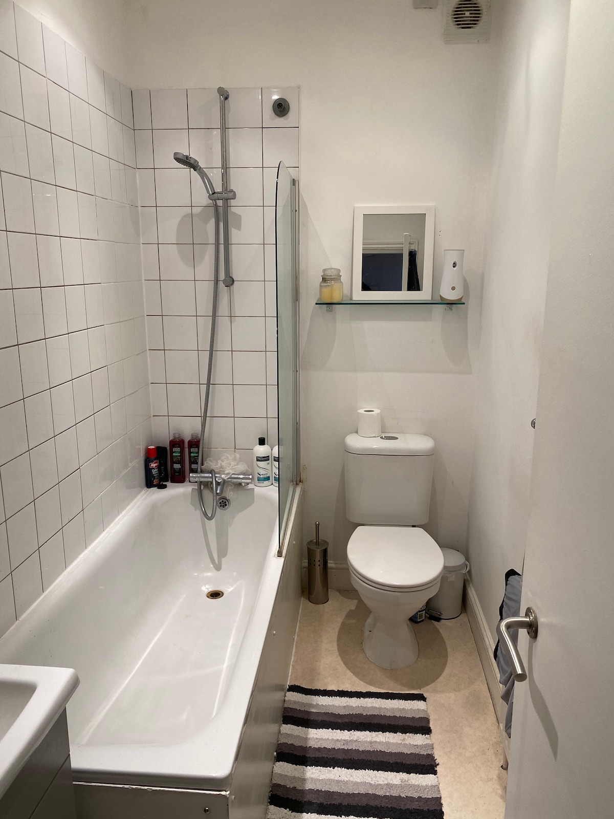 A clean bathroom is shown, featuring a bathtub with an attached shower and glass screen. A small mirror and toiletries are visible on the wall. A striped bath mat lies on the floor, next to a modern toilet and additional wall-mounted storage.