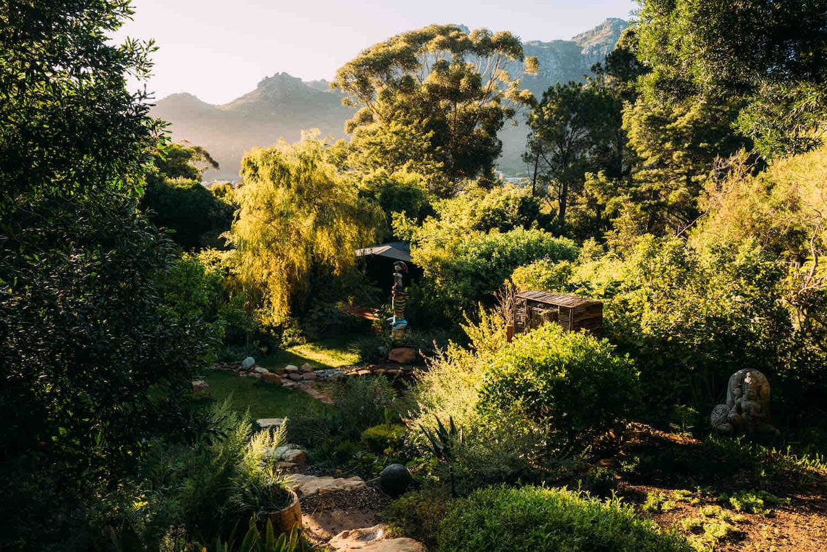 A lush garden is surrounded by diverse greenery, featuring tall trees and varied shrubbery. A serene pathway winds through the landscape, leading to an outdoor seating area shaded by an umbrella. In the background, mountain peaks rise softly against the morning sky.