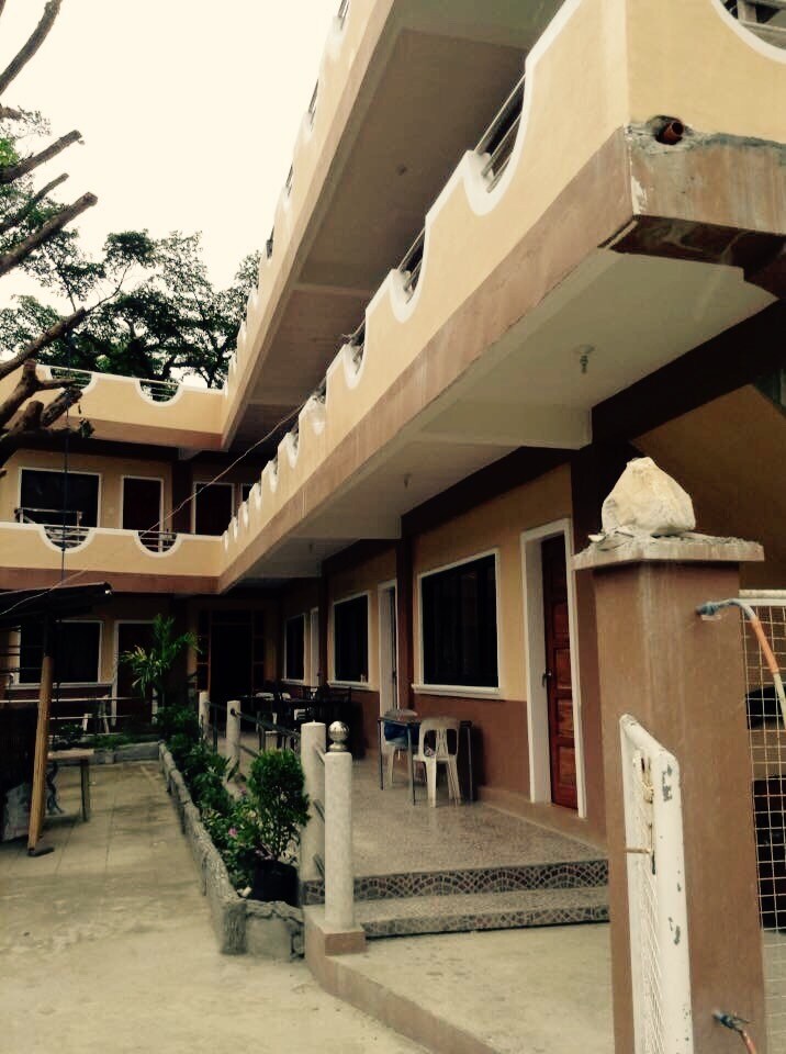 An exterior view of a multi-level building features a spacious balcony with curved railings. The ground level contains a stone pathway leading to the entrance, bordered by greenery. Several outdoor seating options are visible, providing ample space for relaxation.