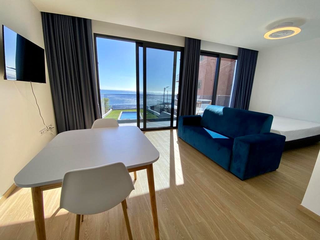 A bright and airy living area features large glass doors that open to an ocean view, allowing natural light to fill the space. A cozy blue sofa is positioned near a small dining table and chairs. Beyond the doors, a glimpse of the garden and pool can be seen.