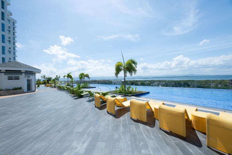 An outdoor pool area is shown, featuring a shimmering infinity edge pool extending towards scenic views. Yellow lounge chairs are positioned along the side, surrounded by greenery. The expansive patio is paved with large tiles, providing ample space for relaxation under a clear blue sky.