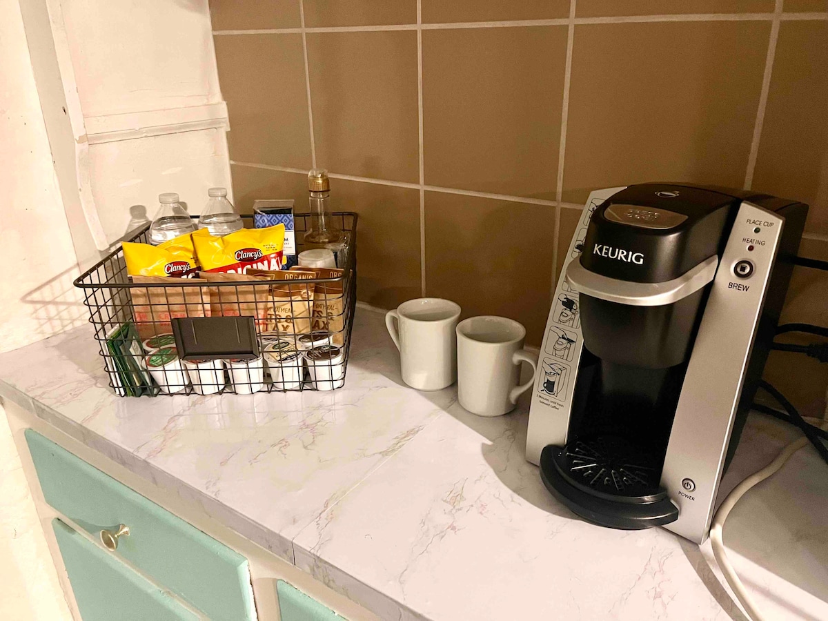 A Keurig coffee maker is positioned on a marble countertop, accompanied by two white cups. A wire basket holds a selection of snacks and bottled water, set against a backdrop of light brown tiled walls.