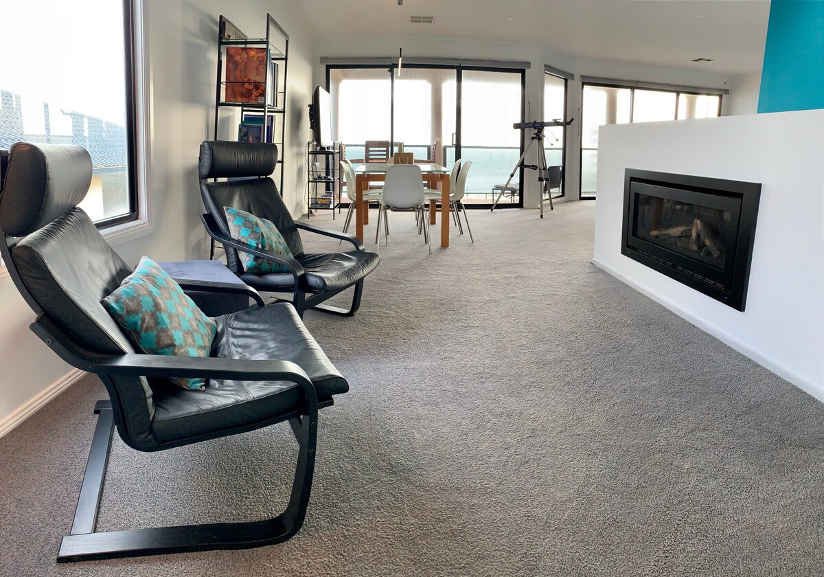A spacious living area features two black lounge chairs with decorative cushions, positioned beside a sleek fireplace. A dining table is visible in the background, complemented by several windows that overlook the scenic views. The room is designed with neutral-colored carpeting.