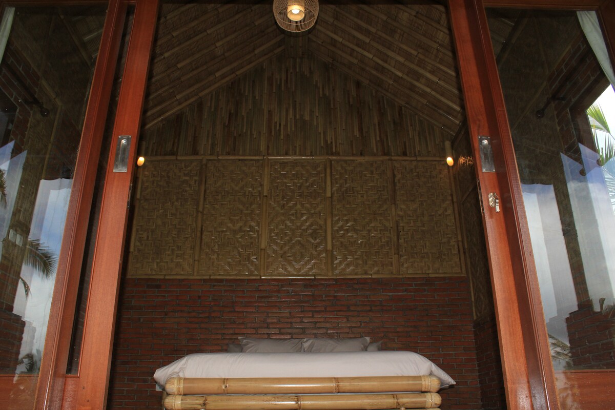 A cozy sleeping area is portrayed with natural bamboo accents, featuring a low-lying bed adorned with neutral bedding. The warm, textured brick wall contrasts with the high, thatched ceiling, while large glass doors allow natural light to brighten the space.