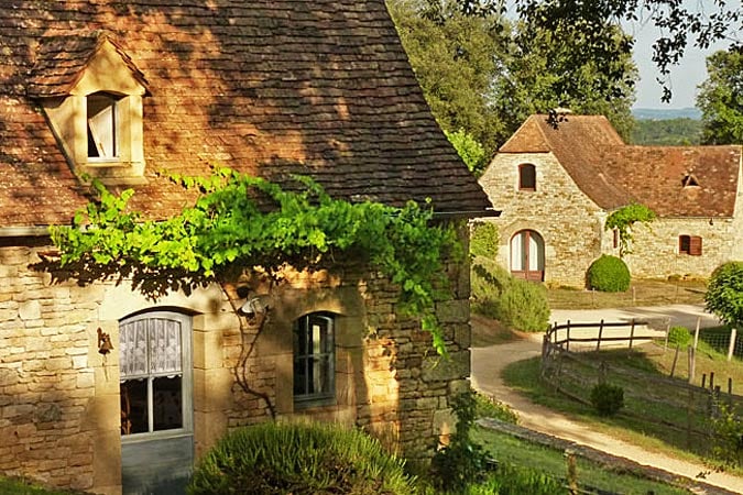 A charming stone cottage features a warm roof and is surrounded by greenery. A pathway leads through the landscape, guiding the eye toward additional rustic buildings in the background. Soft light filters through the trees, enhancing the peaceful ambiance of the setting.
