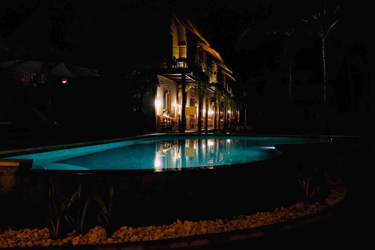 A tranquil pool reflects the softly lit exterior of the bungalows at night. The illuminated building features a welcoming porch and outdoor seating, surrounded by palm trees, enhancing the peaceful ambiance.