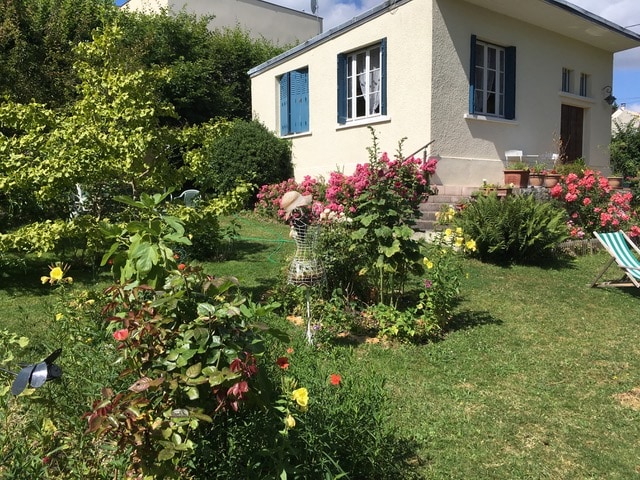 A charming house sits amidst a vibrant garden, featuring lush greenery and colorful flowering plants. The exterior is painted in light tones, with blue shutters framing the windows. A stone pathway leads to the entrance, creating a welcoming approach to the residence.