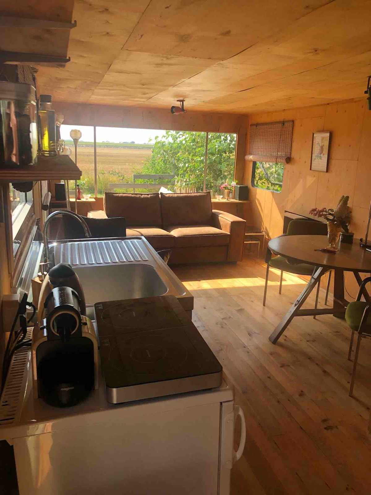 The open-plan living area features large windows offering a view of natural surroundings. A comfortable couch is positioned near the light, while a dining table with chairs stands nearby. A kitchenette with basic appliances is visible, complemented by wooden surfaces that enhance the cozy ambiance.