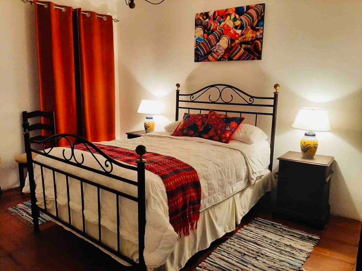 A spacious room features a wrought iron bed adorned with decorative pillows and a vibrant blanket. Two bedside tables with lamps provide soft lighting. Warm orange curtains frame the window, and a colorful textile artwork hangs above the bed, enhancing the room's eclectic design.