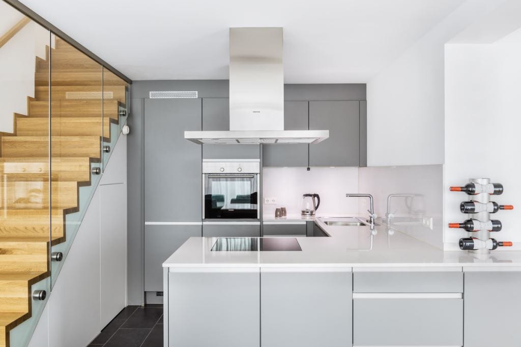 A modern kitchen features sleek gray cabinetry and a central island with a built-in cooktop. Stainless steel appliances, including an oven and a refrigerator, are visible. The back wall is accented by a minimalist backsplash, while a staircase with wooden steps and glass railing adds a contemporary touch.