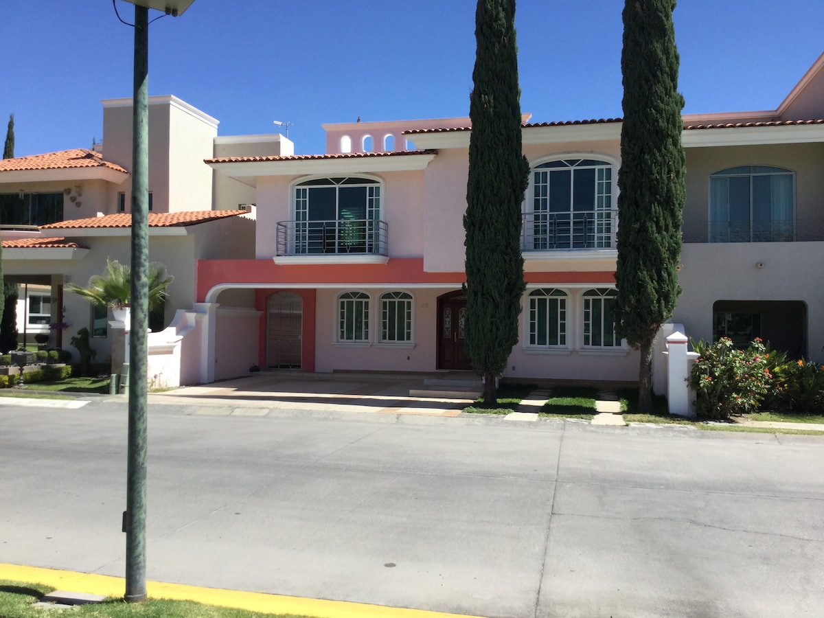 A two-story house is shown, featuring a light pink facade with white accents. Large windows adorn the front, allowing natural light to enter. Tall, green trees flank the sides, and a neatly maintained garden is visible at the front, complementing the inviting appearance of the home.