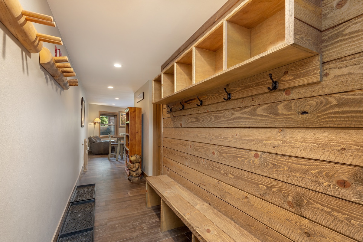 A cozy entryway is highlighted by a wooden bench and storage nooks against a rustic wooden wall. Hooks are available for hanging items. The space leads to a well-lit living area visible in the background, enhancing the inviting ambiance.