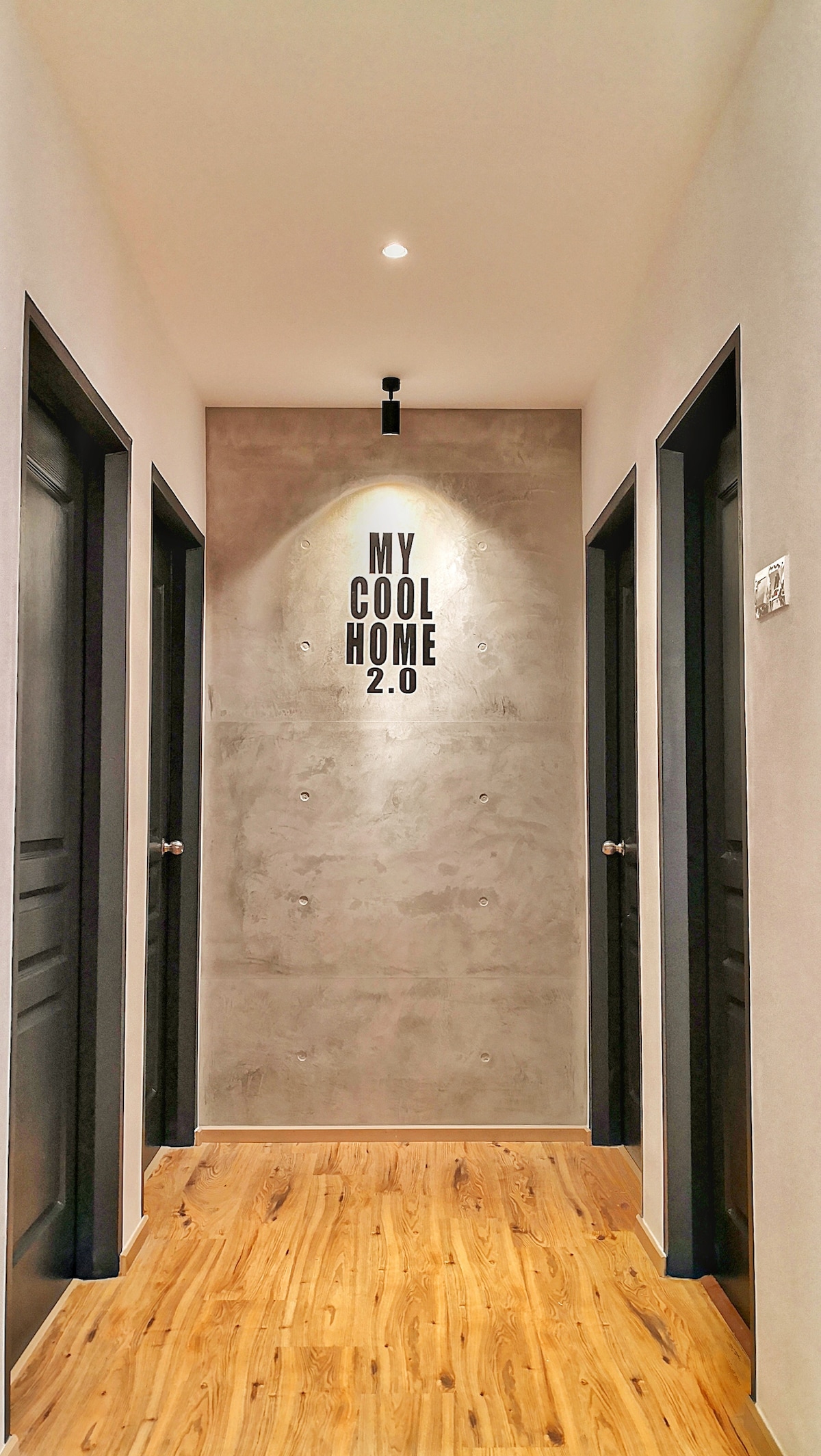 A modern corridor is shown, featuring a cement-textured wall with the text 'MY COOL HOME 2.0' displayed prominently. The hallway is flanked by black doors on either side, and wooden flooring adds warmth to the space. Soft lighting highlights the corridor's contemporary design.