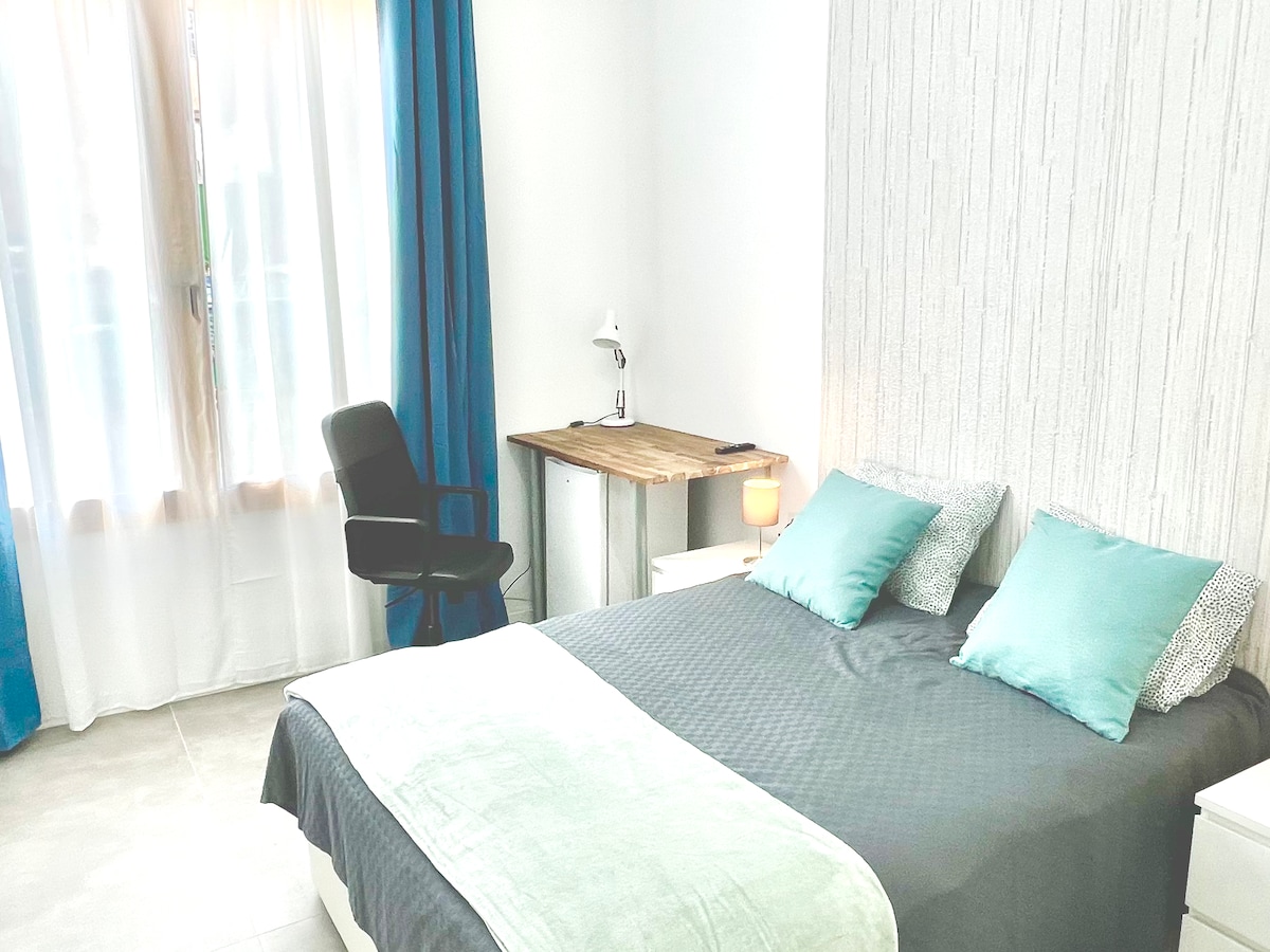 A bright room features a spacious bed dressed in soft bedding. Two accent pillows sit atop the bed. A desk and chair are positioned nearby, illuminated by natural light from a large window with sheer curtains. The walls are adorned with light, neutral tones.