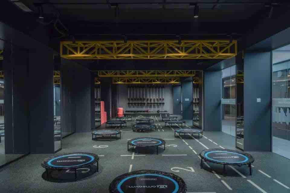 A fitness studio is depicted, featuring multiple circular trampolines arranged on a dark carpeted floor. Bright yellow beams are visible in the ceiling, contrasting with the dark walls. Mirrors line one side of the room, reflecting the energetic setup designed for group exercise activities.
