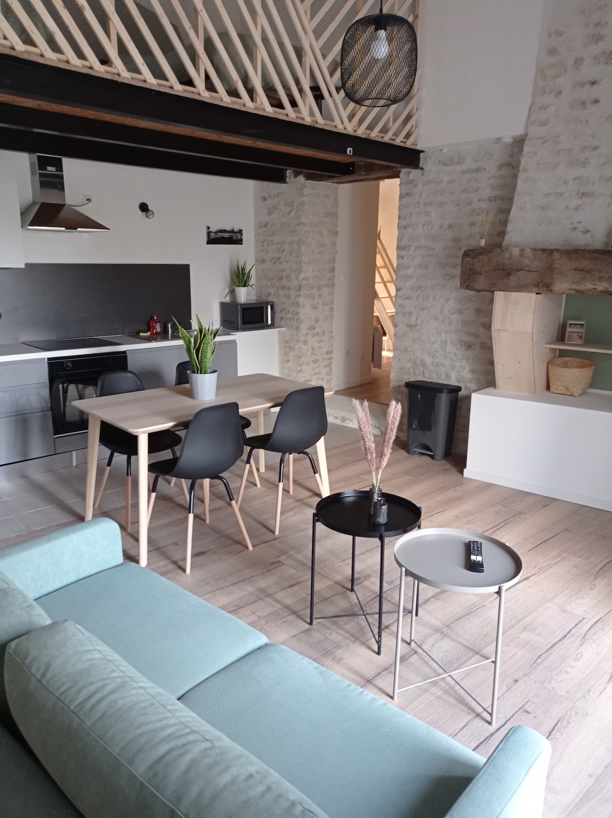 A modern living space features a kitchenette with stainless steel appliances and a dining area with a light wooden table surrounded by four black chairs. In the foreground, a soft sofa sits near two side tables. A large window illuminates the room, enhancing the stone wall's texture.